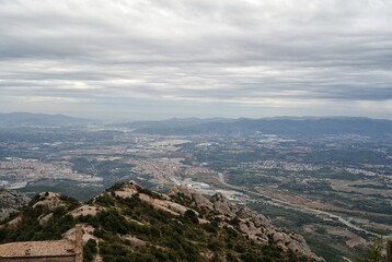 Obraz premium A high angle panoramic view from Montserrat mountain in Catalonia, Spain. Rocky peaks in the foreground, green valley with towns and roads under a grey cloudy sky during a daytime in the summer