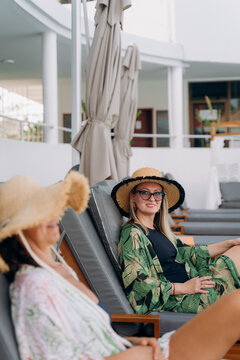 Two women sit in lounge chairs at a resort pool, wearing large hats and casual outfits. They appear to enjoy the warm weather and their time together under umbrellas.
