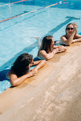 Three women relax and talk at the side of a swimming pool. They are wearing swimsuits and enjoying their time together in the water under clear blue skies.