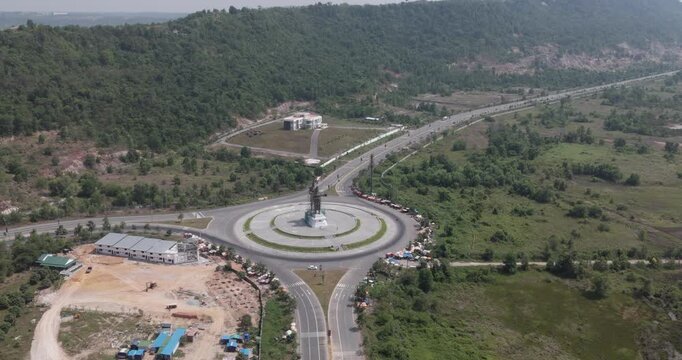 aerial turning view of the Preah Thong and Neang Neang statue at a major roundabout in Sihanoukville, Cambodia, featuring circular traffic flow, roadside markets, and lush greenery.