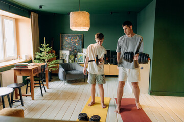 Two individuals focus on lifting dumbbells in a home workout area. The space has natural light and is furnished with a table and a chair. © m-art