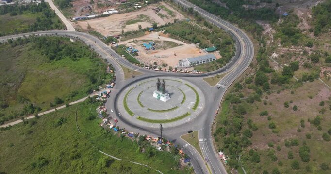 aerial turning view of the Preah Thong and Neang Neang statue at a major roundabout in Sihanoukville, Cambodia, featuring circular traffic flow, roadside markets, and lush greenery.