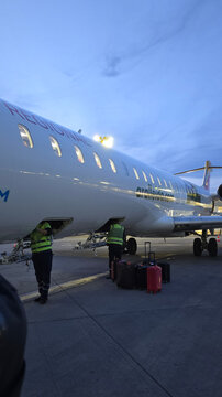 Ground crew members handling passenger baggage on the tarmac next to a parked aircraft during evening operations