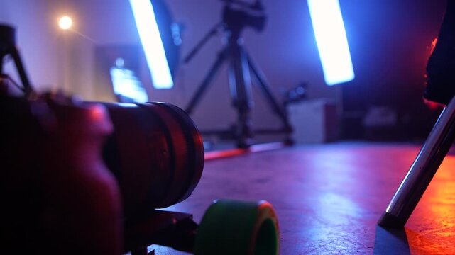 Professional camera mounted on a small dolly with blue and warm stage lights in the background. Concept of filmmaking, video production, backstage technology, media, and creative industry.