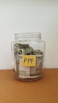 Close up of hand putting india rupee currency note inside transparent glass jar isolated on table with PPF word or text label. Investing money in PPF (Public Provident Fund) retirement scheme concept.