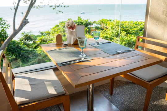 Table set for two with blue water glasses and silver cutlery on a wooden patio overlooking the ocean. Ideal for luxury seaside dining, beach cafe, and summer vacation lifestyle themes.
