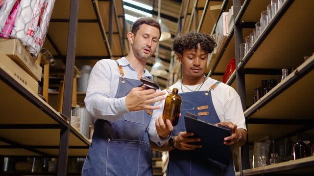 Diverse male colleagues wearing denim aprons inspect a brown spray bottle while checking inventory lists in a warehouse. Professional teamwork and small business product management in a stockroom.