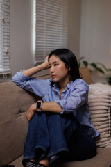 Obraz premium Asian woman thinking while sitting alone on a sofa, looking sad and experiencing stress or loneliness, representing emotional pain and mental health issues