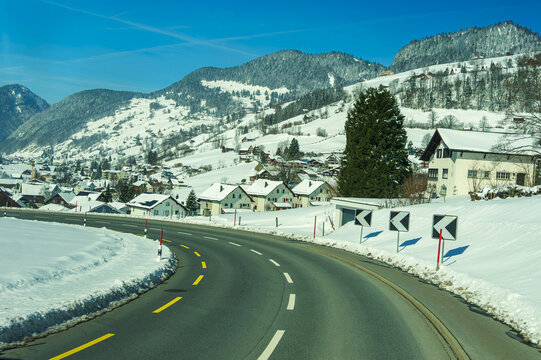Snowy mountain village with winding road, clear blue sky, and scattered houses. serene winter landscape features snow covered rooftops and distant forested hills, creating peaceful atmosphere
