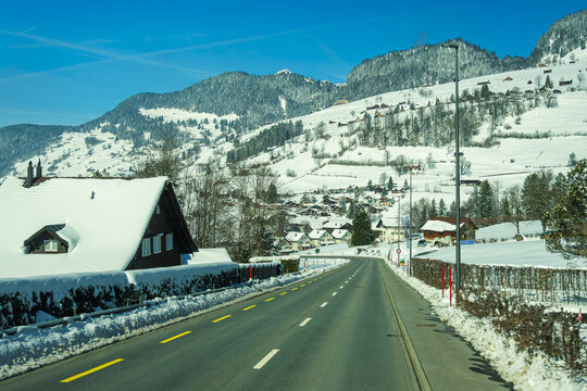 Snow covered road leading to picturesque village nestled in mountains, with charming houses and clear blue sky. serene winter landscape evokes sense of tranquility and beauty