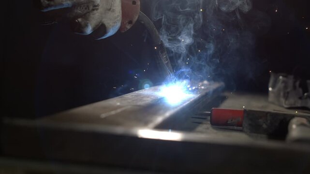 A welder constructs a metal piece in a workshop at night. Bright sparks and smoke are seen as the welding machine fuses the metal together. Tools and materials are nearby.