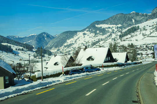 Snow covered alpine village with traditional houses and winding road under clear blue sky, surrounded by majestic mountains. serene winter landscape evokes sense of tranquility