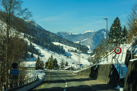 Snowy mountain road with clear blue sky, surrounded by trees and hills, creating serene winter landscape. speed limit sign is visible, indicating 50 km/h