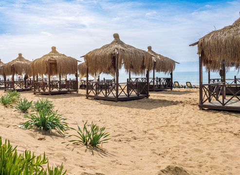 Traditional wooden beach pavilions with thatched straw roofs on sandy beach in Belek, Turkey. Luxury resort scene, calm Mediterranean sea under blue sky, summer vacation and high-end travel concepts.