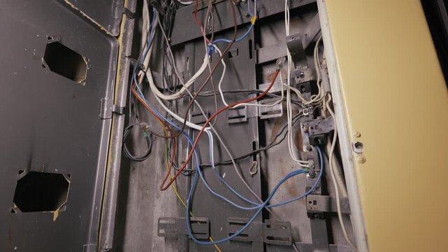 Old electrical panel displays chaotic tangle of exposed wires and dust. Camera tilts up revealing rusty metal components and neglected connections inside open cabinet.