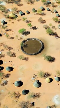 Aerial drone view of a traditional circular Himba village settlement in northern Namibia with huts arranged around a central livestock enclosure. Vertical