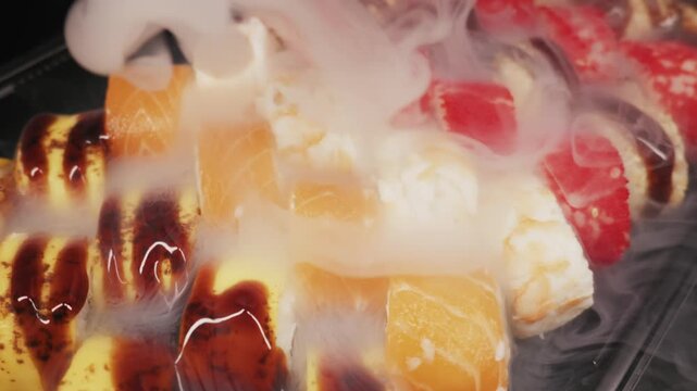 Various sushi types including salmon, eel, and tobiko rolls sit on a black platter. Thick white dry ice fog flows over the fresh seafood appetizers.