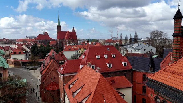 Cinematic drone flight over Ostrow Tumski in Wroclaw, showing an aerial panorama of the old town, red tiled roofs, Holy Cross Church tower, and historic gothic architecture under a cloudy sky.