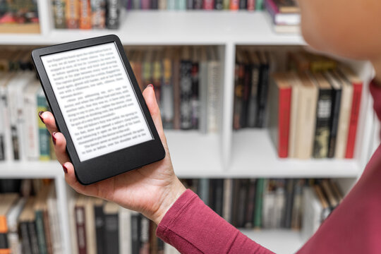 a woman holds an e-book in her hand. a woman's hand holding an e-book. A woman reads an e-reader near a bookcase. an e-book against the background of paper books. A woman reading an e-book at home