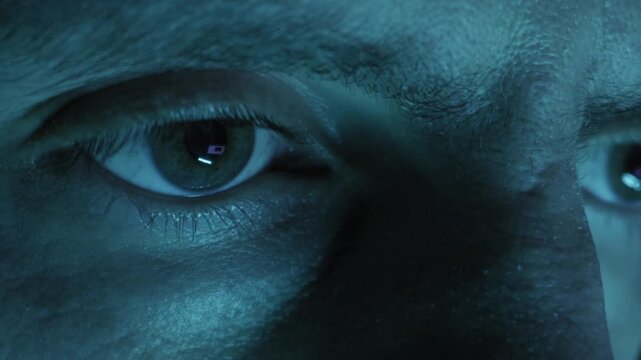 Close-up of a man intense eyes staring at a monitor in a dark room. The screen reflects in his pupils and illuminates his face. Concept of technology, hacking, focus, and surveillance.