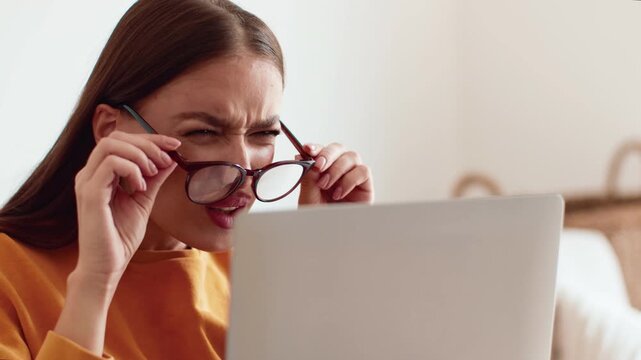 A woman with light skin sits at a table, looking intently at her laptop. She adjusts her glasses while displaying concern. The setting appears to be a cozy indoor space.