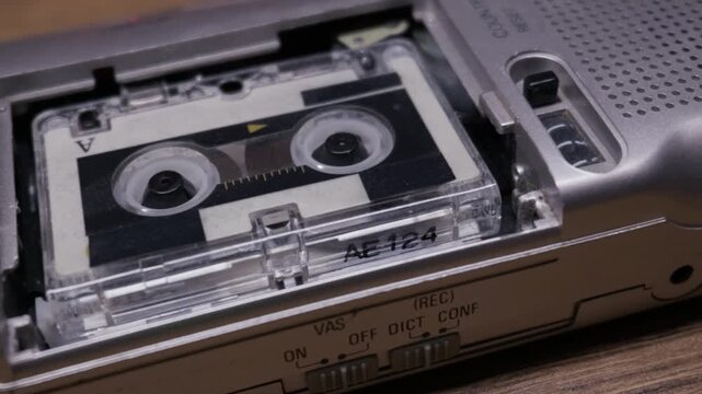 A silver vintage microcassette recorder operates on a wooden desk with the tape spinning inside. Concept of retro journalism, espionage technology, and analog audio history.