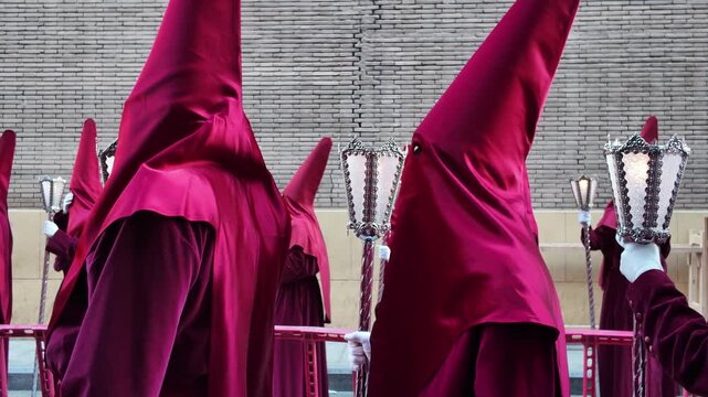 Group of nazarenos penitents dressed in deep red burgundy robes and tall pointed capirote hoods carrying lanterns during solemn Semana Santa Holy Week Catholic procession Spain Murcia