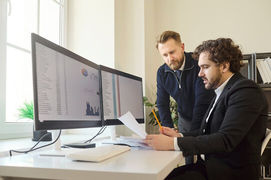 Employees colleagues use office computers for analysis discussion. Two professionals compare charts on dual monitors and planning from a report. Teamwork and focused productivity concept.