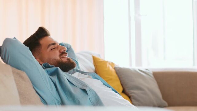 A man with a beard smiles while relaxing on a couch. He leans back with his arms behind his head. The room is bright with soft light coming through the window.