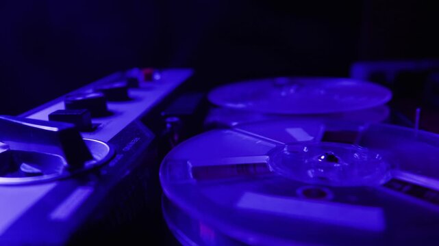 A vintage reel to reel tape recorder operates in a dark room. Transparent plastic spools spin as the magnetic audio tape plays. Neon lighting shifts through various bright colors.