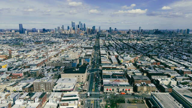 Long axis city sweep along Broad St highlights uninterrupted neighborhood grids extending toward downtown towers. Light roof membranes, muted masonry, bright midday clarity define Philly, PA