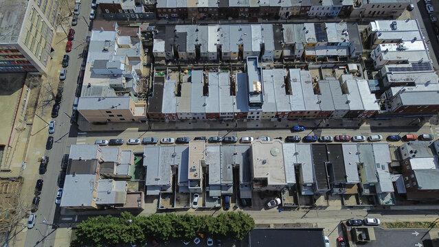 Courtyard edge row blocks near S 11th St and Tasker St display compact housing patterns beside dense modern institutional structure. Light roof membranes, subtle masonry shifts in Philly, PA