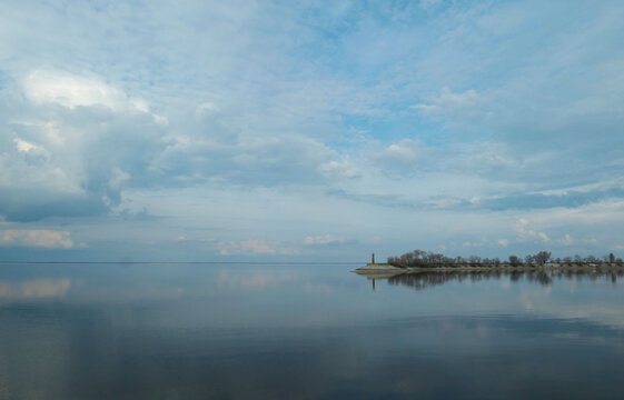 Serene panoramic view of a wide river or lake with perfectly smooth water surface reflecting the cloudy sky. In the distance, a narrow sandspit with a lighthouse and a row of trees. Perfect background