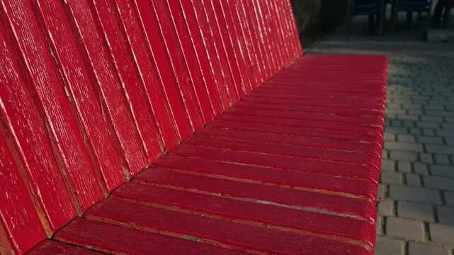Bright red wooden park bench shows weathered texture and peeling paint under strong warm sunlight. Deep shadows fall between the parallel slats to create bold visual contrast.