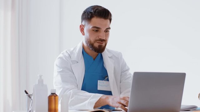 A young man with a beard wearing a white lab coat and blue scrubs sits at a desk in a medical office. He focuses on his laptop while surrounded by medical supplies and tools.