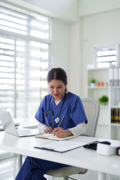 Asian adult female healthcare professional writes patient notes medical report using laptop computer modern clinic office setting health technology