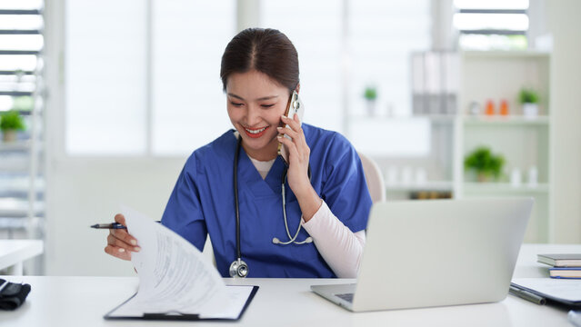Asian adult healthcare professional multitask communication client patient on phone reviewing document using laptop medical clinic office