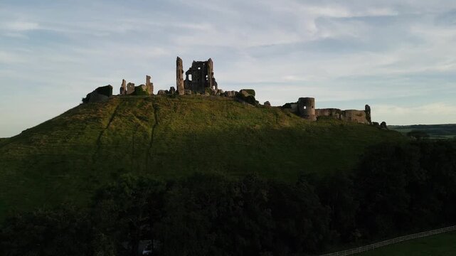 Corfe Castle ruins on top of the hill at sunset. Aerial footage flying above the trees towards the castle. Historic medieval site, popular tourist attraction in Dorset, UK.