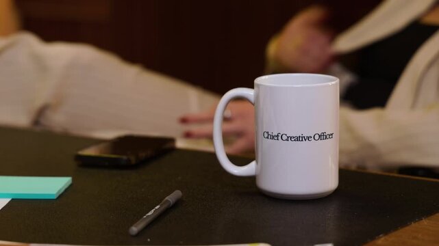White coffee mug with Chief Creative Officer text on office desk as a businesswoman grabs their phone and puts their feet up on the desk.