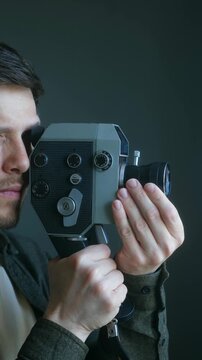 Focused cameraman looking through viewfinder of vintage super 8 camera while recording, his hands adjusting lens and holding grip against dark, solid background. Cameraman filming with vintage camera