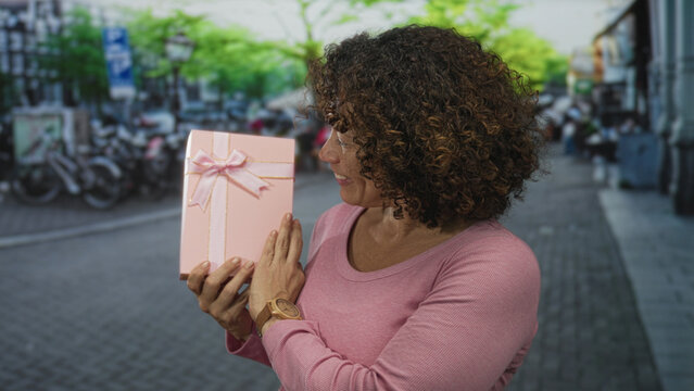Woman smiles and holds pink giftbox on a sunny colorful leafy bustling street; gratitude celebration joy.
