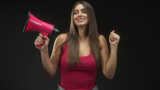 Woman holding pink megaphone with raised fist in studio, smiling in sleeveless tanktop; confidence empowerment activism.