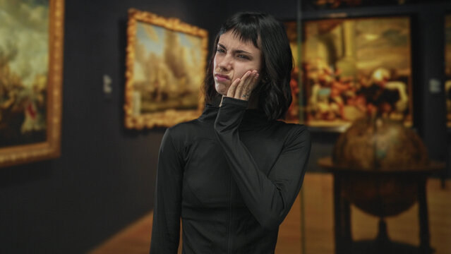 Woman holds hand on cheek while standing before large framed paintings in modern gallery building; pain.
