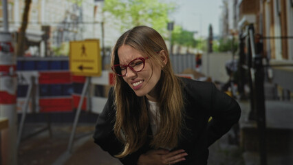 Fototapeta premium Woman clutching stomach with hand and wincing on street near barricade; distress pain urgency emergency.