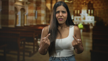 Fototapeta premium Woman points fingers to chest in church aisle wearing white tank and jeans with puzzled expression and raised brows; confusion doubt.
