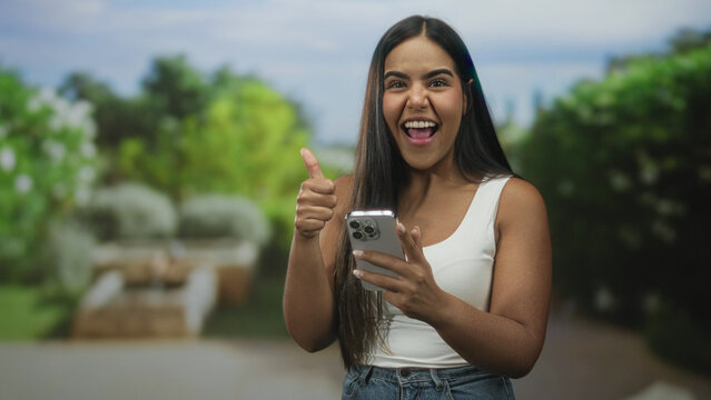 Young hispanic woman holding smartphone and giving thumbs up while looking at screen in studio with blurred garden backdrop; approval.