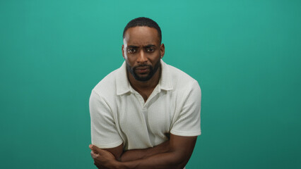 Fototapeta premium Man with crossed arms showing forearms and white polo shirt in teal studio three quarter closeup pose leaning slightly forward and facing camera; concern introspection.