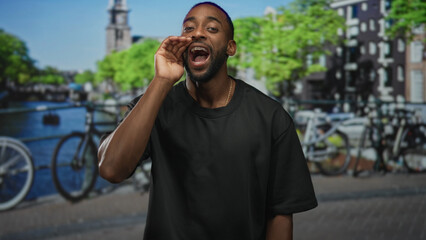 Fototapeta premium Man cupping hand to mouth and clasping hands while smiling on a canal street in amsterdam with bicycles by the water; joy camaraderie.
