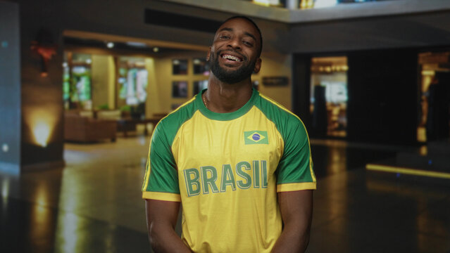 Man smiling, standing in a hotel building lobby wearing a yellow and green brazil jersey with flag patch, facing camera; pride.