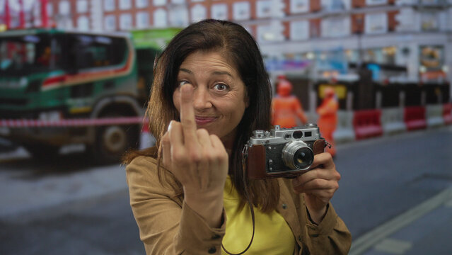 Woman holding vintage camera making middlefinger gesture in front of building; bold defiance attitude.